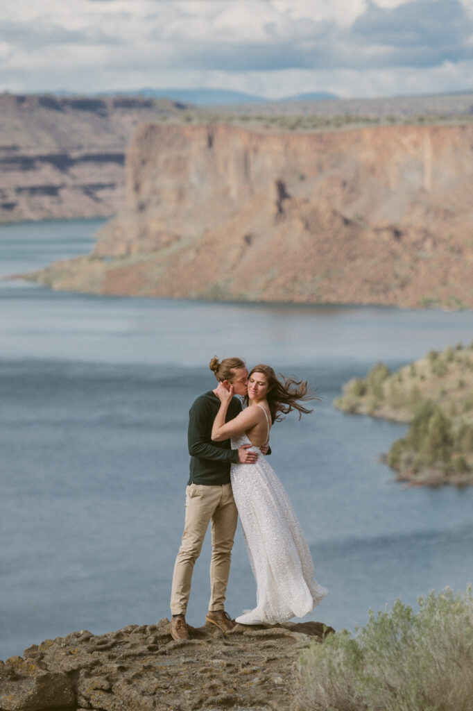 A man and woman stand at an overlook on the Tam-a-lau Trail in the Cove Palisades State Park in Oregon. The woman's hair is whipping in the wind. She has her face turned away from the man as he holds her waist and kisses her cheek.