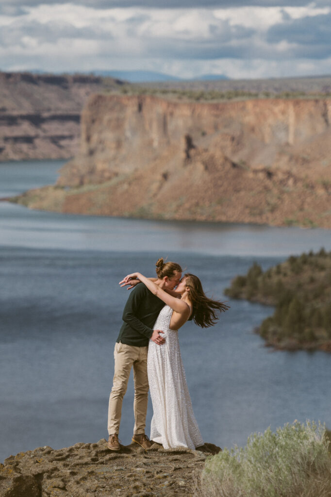 A man and woman stand at an overlook on the Tam-a-lau Trail in the Cove Palisades State Park in Oregon. The woman's hair is whipping in the wind. She has both hands crossed at the wrist behind the man's head. He is holding her waist and kissing her on the lips.