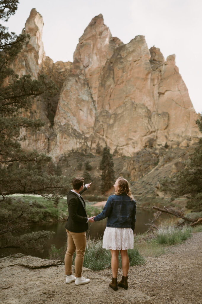 Two women hold hands as they look away from the camera in Smith Rock State Park, Oregon. One woman is wearing a white sun dress with denim jacket while the other woman is wearing khaki pants, a button down shirt, tweed vest, and jacket. The woman wearing the khaki pants is pointing towards a distant rock climber.