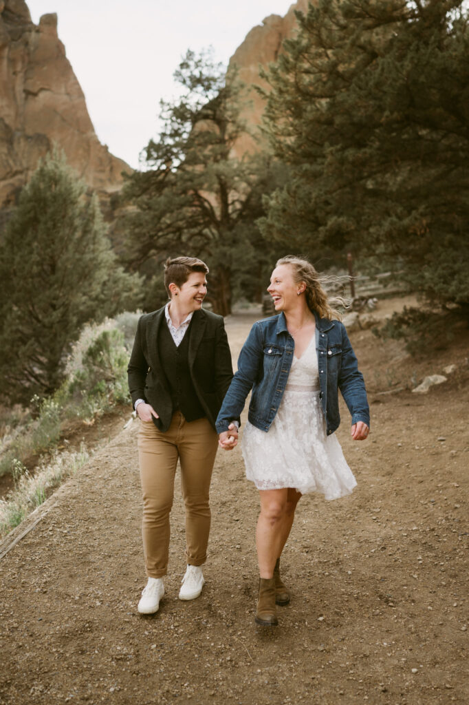 Two women hold hands as they walk towards the camera in Smith Rock State Park, Oregon. One woman is wearing a white sun dress with denim jacket while the other woman is wearing khaki pants, a button down shirt, tweed vest, and jacket.