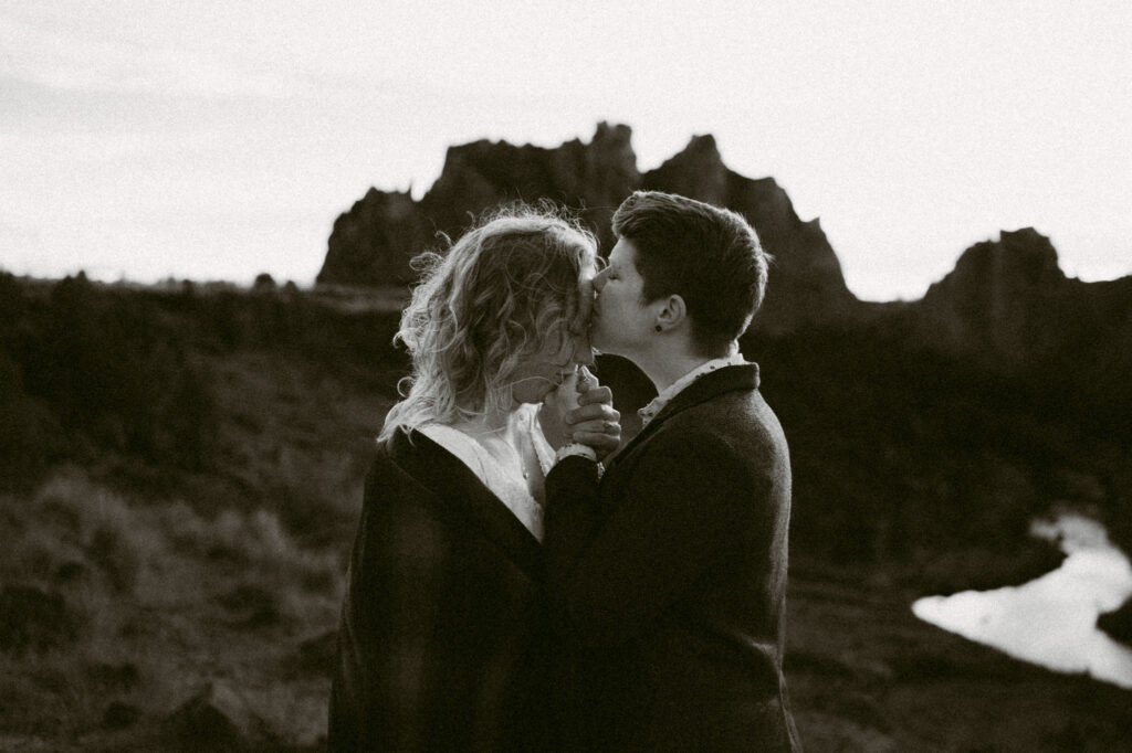 A black and white image of two women embracing at Smith Rock State Park. They're holding hands between their two bodies. One woman is kissing their fingers while the second woman is kissing the first woman's forehead.