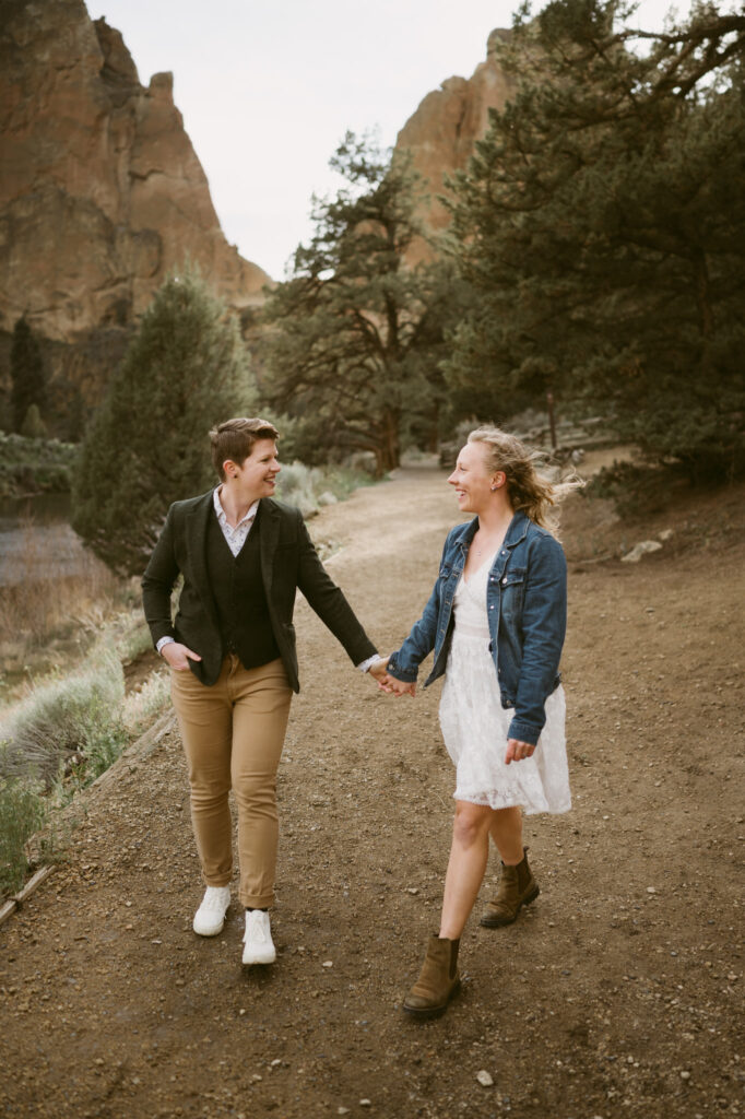 Two women hold hands as they walk towards the camera in Smith Rock State Park, Oregon. One woman is wearing a white sun dress with denim jacket while the other woman is wearing khaki pants, a button down shirt, tweed vest, and jacket.