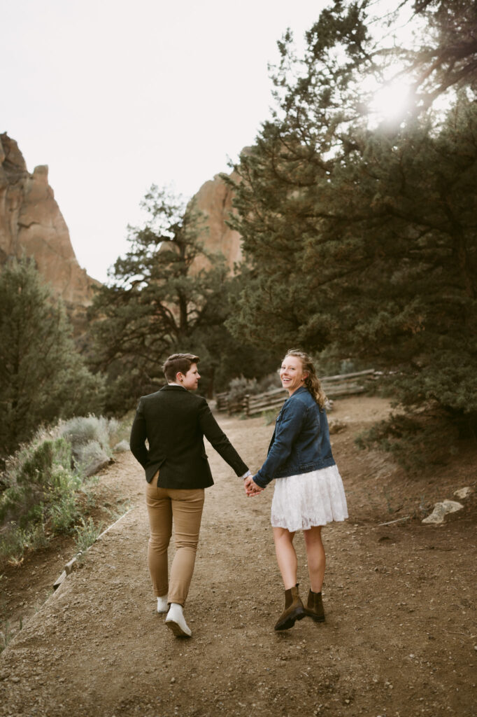Two women hold hands as they walk away from the camera in Smith Rock State Park, Oregon. One woman is wearing a white sun dress with denim jacket while the other woman is wearing khaki pants, a button down shirt, tweed vest, and jacket. The woman in the dress is glancing over her shoulder, towards the camera.