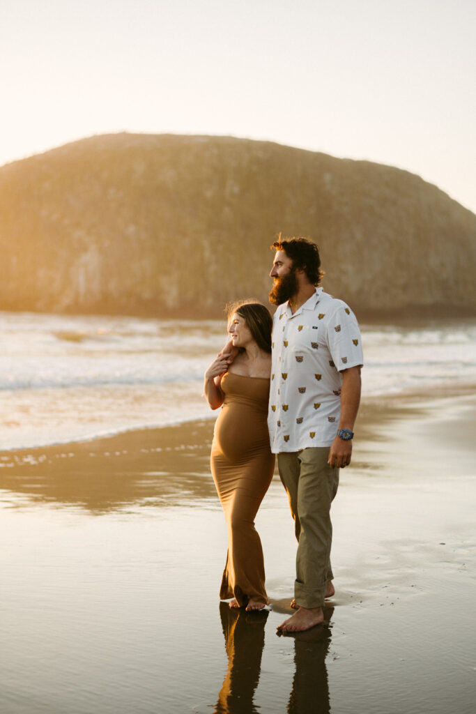 A man drapes his arm over the shoulders of his pregnant wife as they walk down the beach at Seal Rock Recreation Site for their maternity photos.