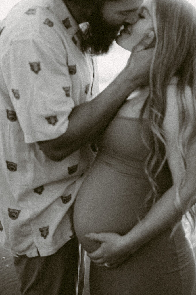 A black and white image of a man cradling his wife's face, kissing her, as she cradles her pregnant belly.