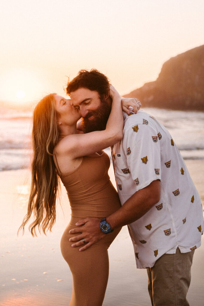 A pregnant woman wraps her arms around her husband's neck and kisses him on the cheek at their maternity session at Seal Rock Recreation Site in Seal Rock, Oregon.