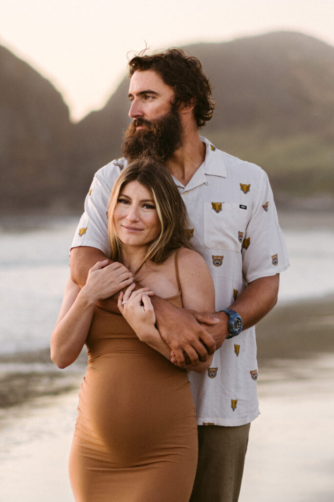 A pregnant woman smiles at the camera as her husband kisses the top of her head at their maternity session at Seal Rock Recreation Site in Seal Rock, Oregon.