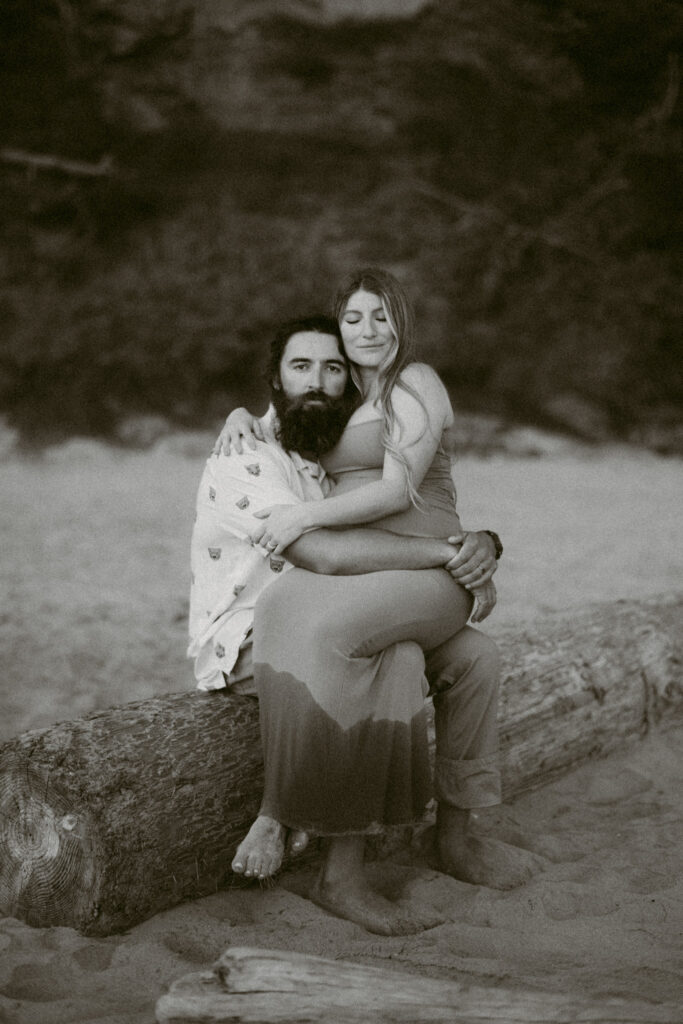 A black and white image of a young couple embracing on a log during their maternity session at Seal Rock Beach in Seal Rock, Oregon.