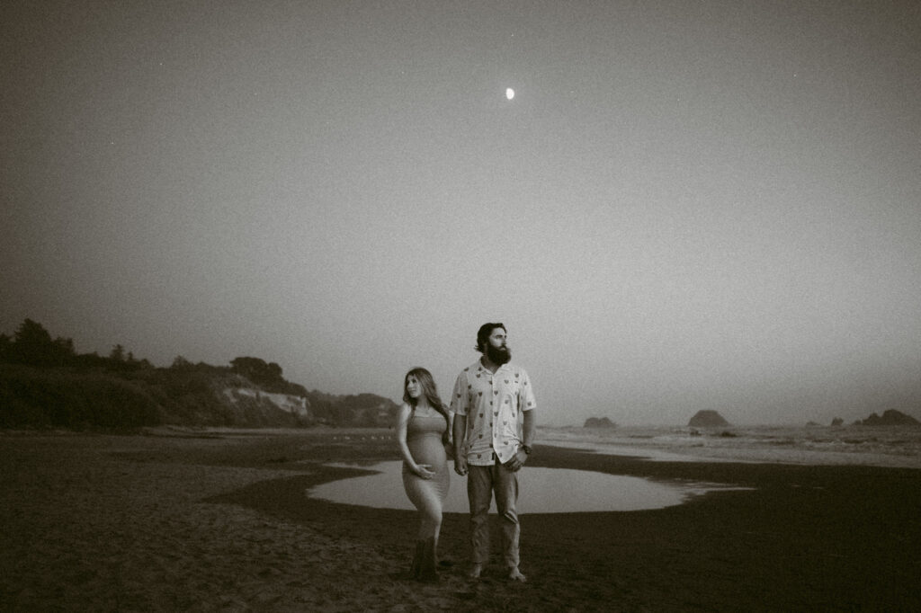 A black and white image of a young couple posing at twilight on Seal Rock Beach during their engagement session in Seal Rock, Oregon.