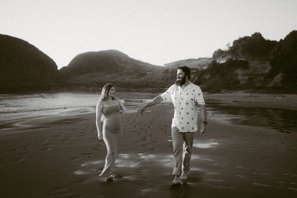 A black and white image of a pregnant woman holding hands with a man as they walk down Seal Rock Beach in Seal Rock, Oregon.