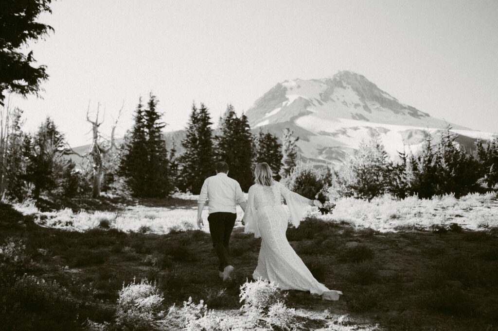 A black and white image of a bride and groom walking towards the summit of Mount Hood during their August wedding at Mount Hood Meadows