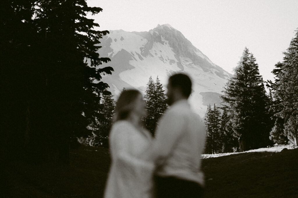 A black and white image showing the summit of Mount Hood. An out-of-focus couple is standing in the foreground.