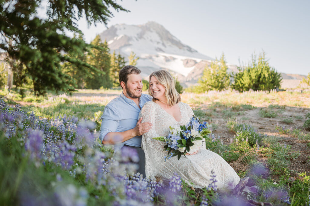 A bride and groom sit amongst a field of lupines during their elopement at Mount Hood Meadows. The summit of Mount Hood looms in the background.