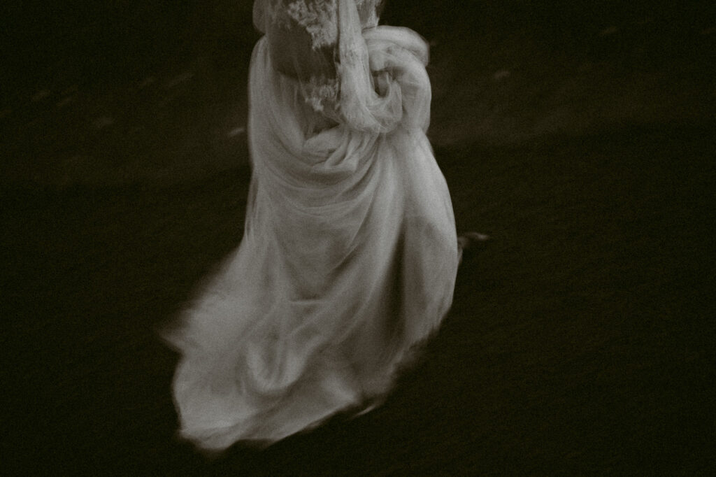A blurry black and white photo depicting a woman holding the train of her wedding dress as she walks by the camera.