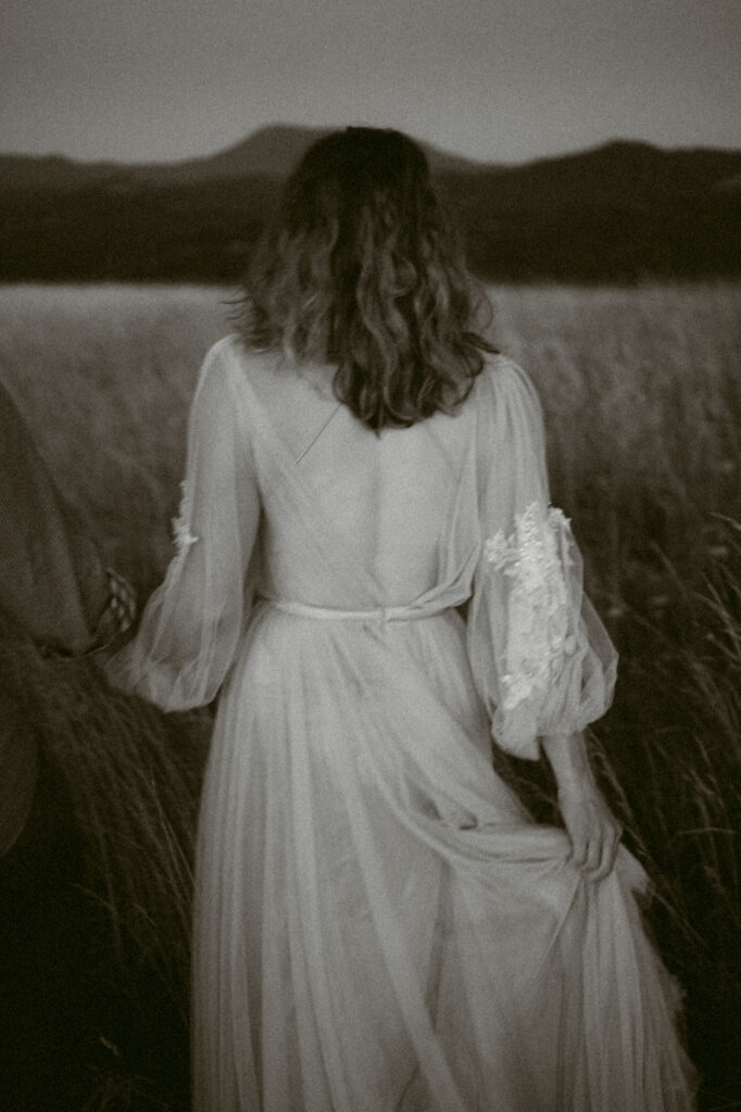 A black and white image of a woman walking away from the camera wearing a wedding dress. Mary's Peak can be seen in the background.