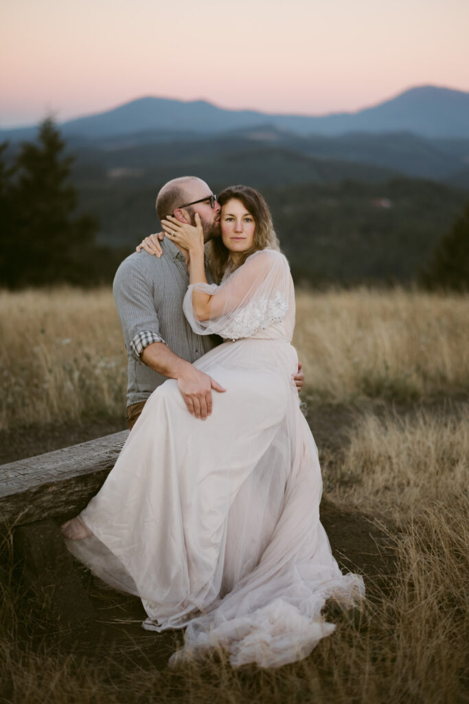 Oregon's Coast Range makes the perfect backdrop for this couple's anniversary photos at Fitton Green in Corvallis. The couple sits on a bench together, with the woman on the man's lap. The man is kissing her cheek while the woman gazes at the camera.