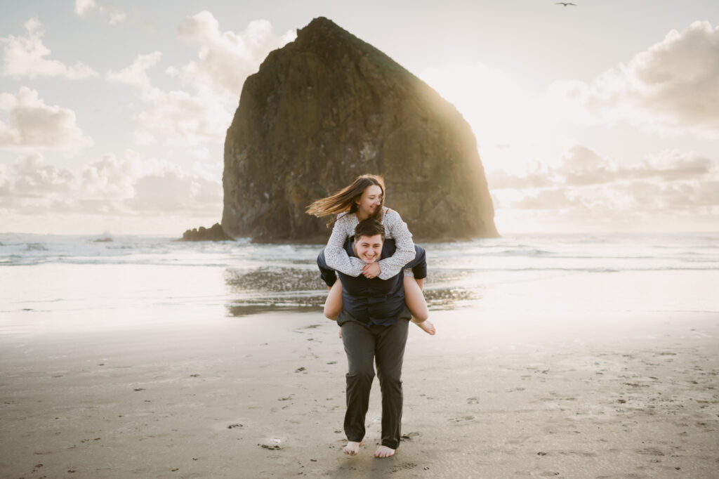 A young couple get playful during their sunset engagement session at Cannon Beach. The man is giving a piggy-back ride to the woman with Haystack Rock in the background.