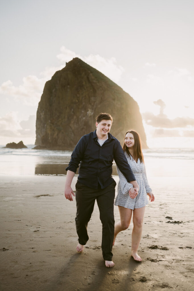 A young couple walks hand in hand down Cannon Beach for their sunset engagement session. Haystack Rock can be seen in the background.