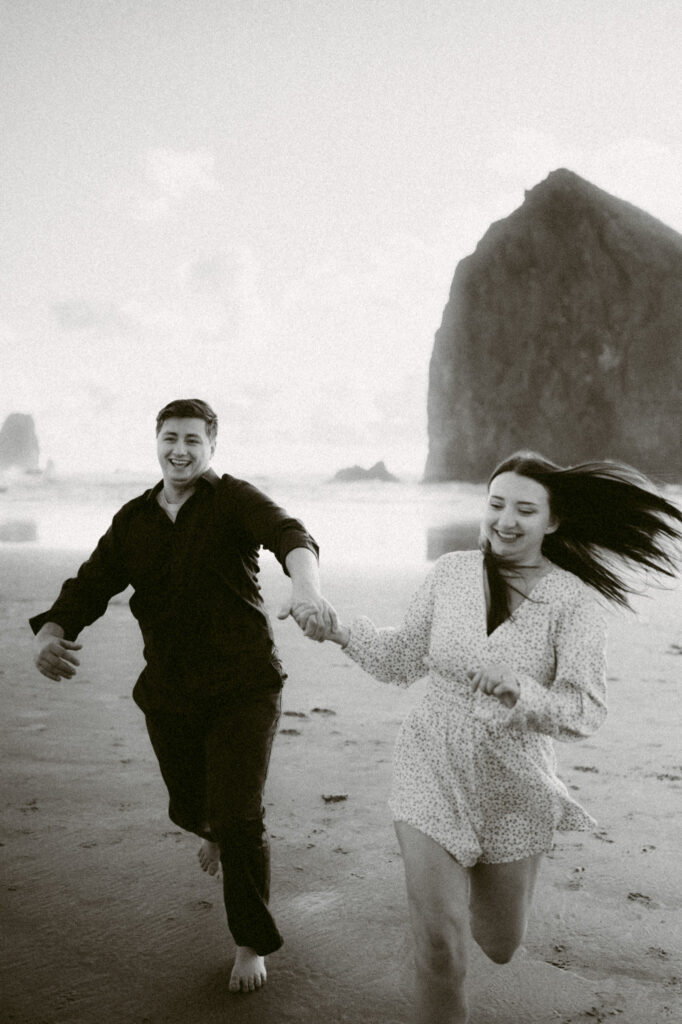 A black and white image of a young couple running hand-in-hand down Cannon Beach.