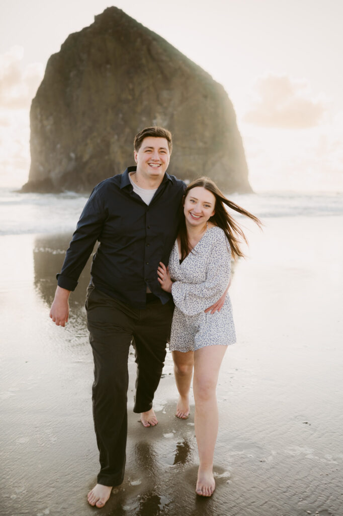 A young couple with their arms wrapped around one another's waists smile as they walk towards the camera. Haystack Rock is visible in the background as they walk along Cannon Beach.