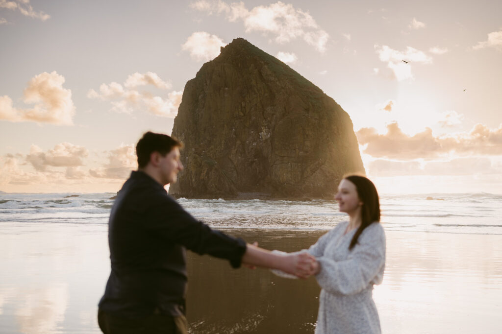 A back-focused photo of Haystack Rock at Cannon Beach during sunset. A young couple stands holding hands in the foreground.