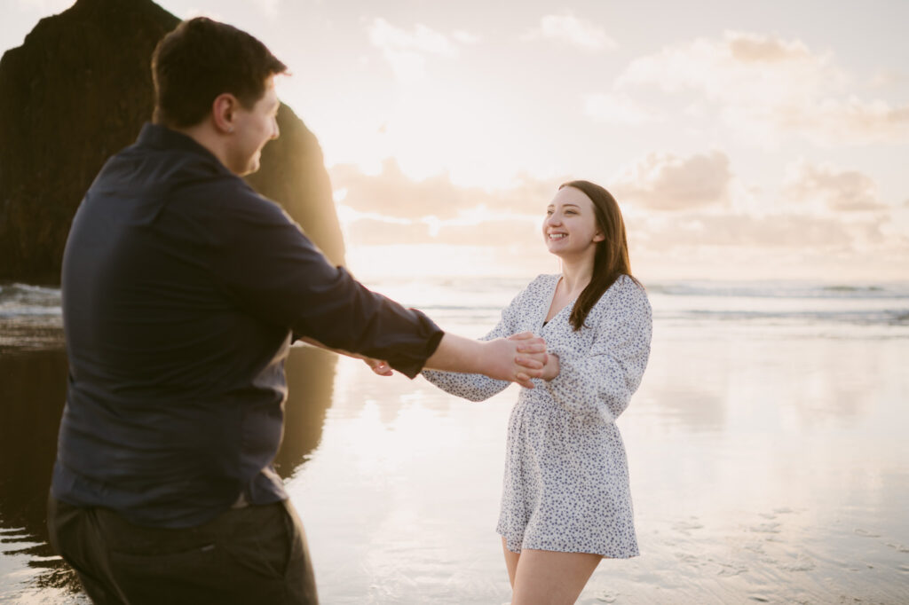 A young couple holds hands and swings around in a circle on Cannon Beach during their engagement session.