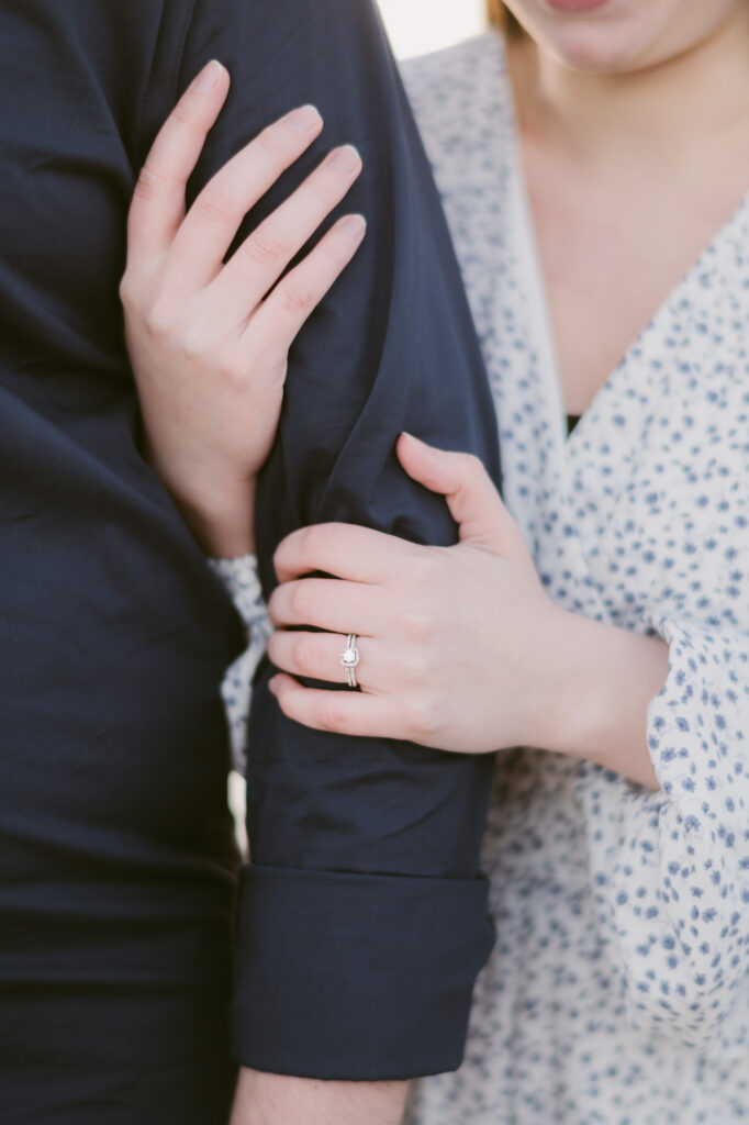 A close-up photo of a woman's hands as she holds onto a man's arm. Her diamond engagement ring is visible.