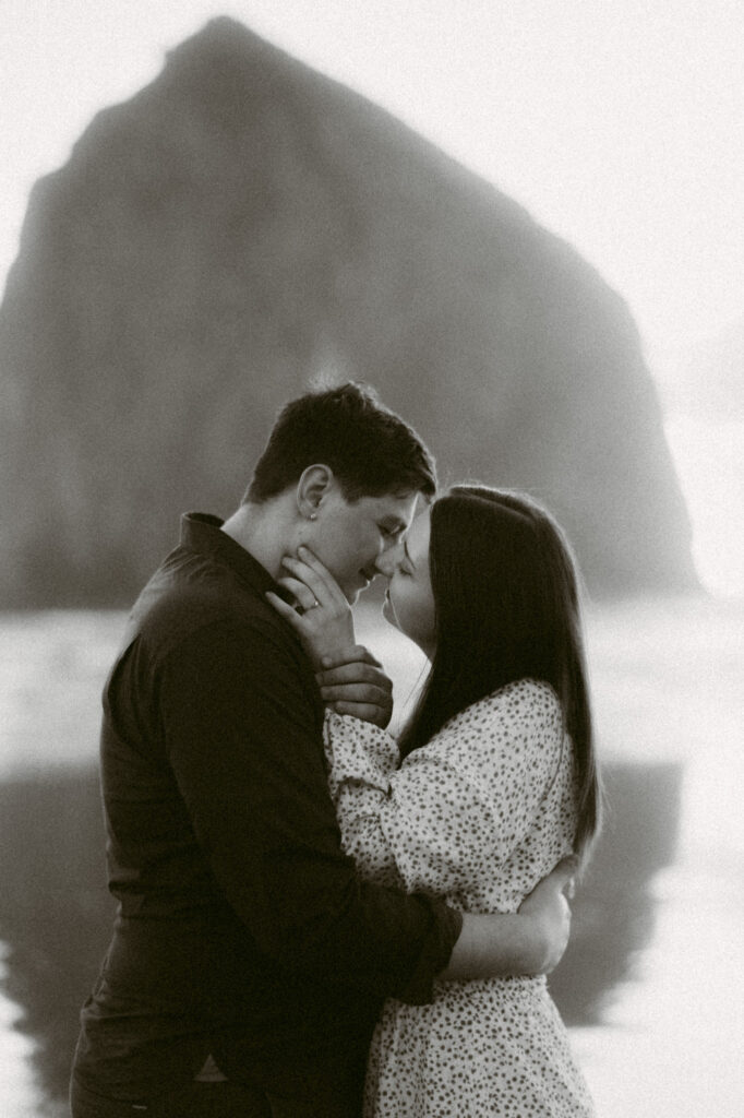 A black and white image of a young couple kissing in front of Haystack Rock at Cannon Beach.