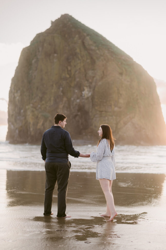 A young couple holds hands and faces away from the camera during their late summer engagement session at Cannon Beach in Oregon.