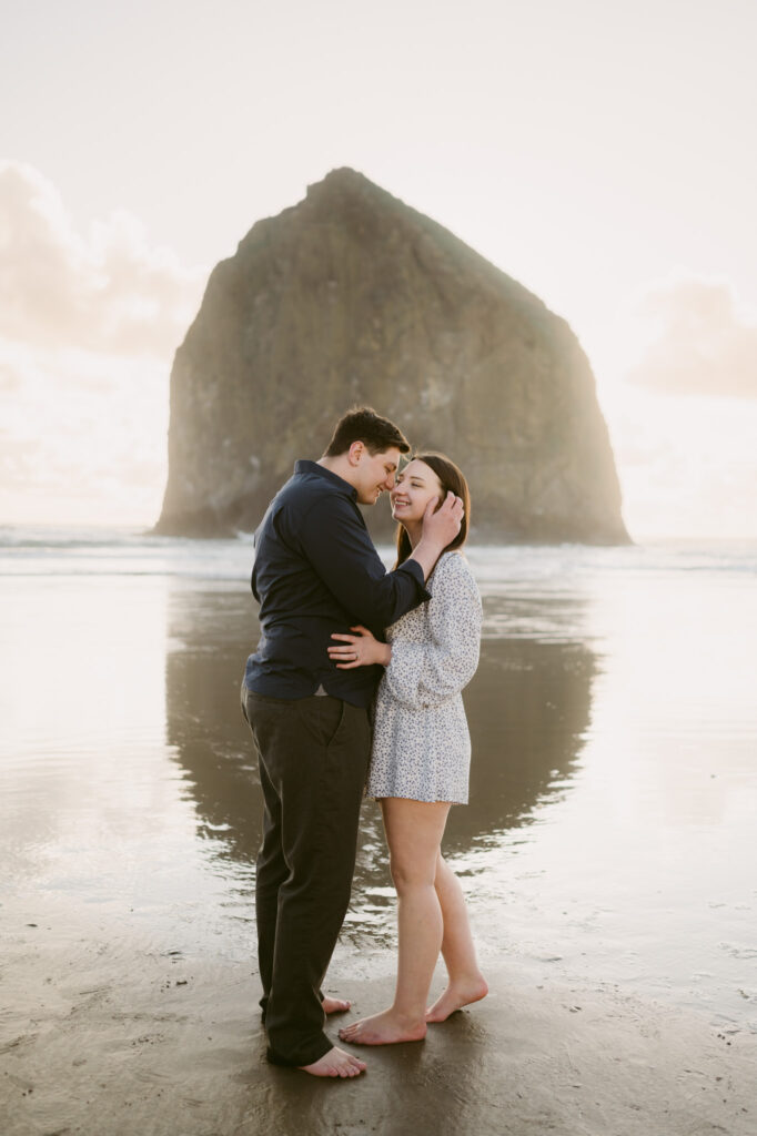 A young couple stops to kiss on Cannon Beach during their engagement session. Haystack Rock stands visible in the background.
