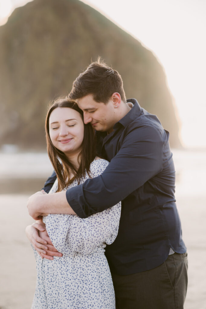 A young couple pose for their engagement photos at Cannon Beach in Oregon. Haystack Rock stands visible in the background as the young man envelopes his fiance in a bear hug.