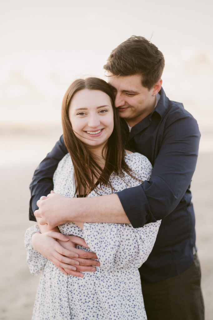 A young heterosexual couple pose for their engagement session at Cannon Beach in Oregon. The man's arms are wrapped around the woman. She's smiling at the camera as the sun sets behind her.
