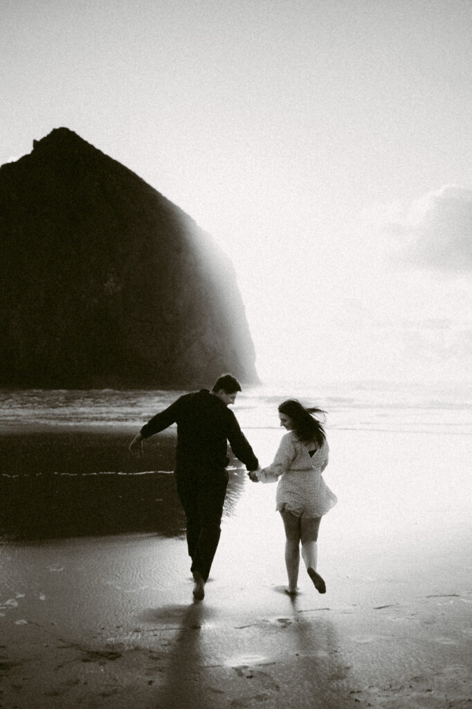 A black and white image from an engagement session at Cannon Beach in Oregon. A young couple holds hands as they run towards Haystack Rock.