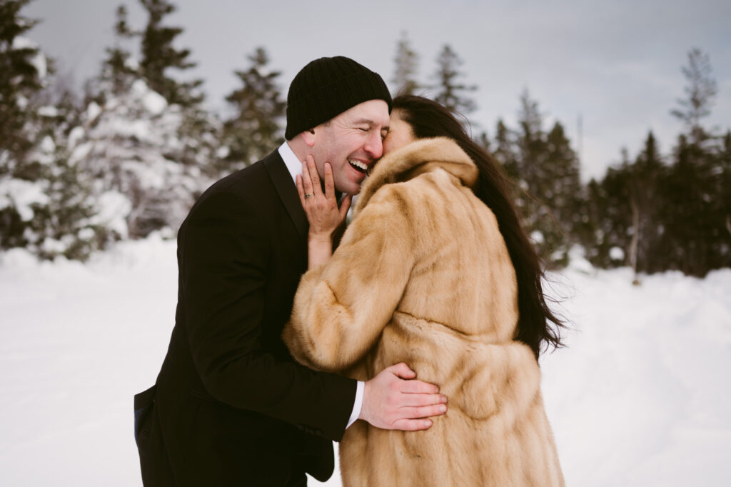 A bride kisses the groom on the cheek amongst a snowy winter scene during their winter wedding in the Adirondacks.