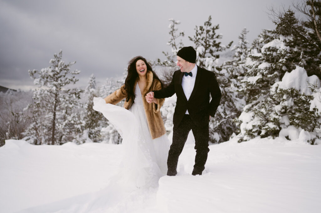 A bride and groom hold hands and laugh as they snowshoe through deep snow during their winter elopement at Cobble Lookout.