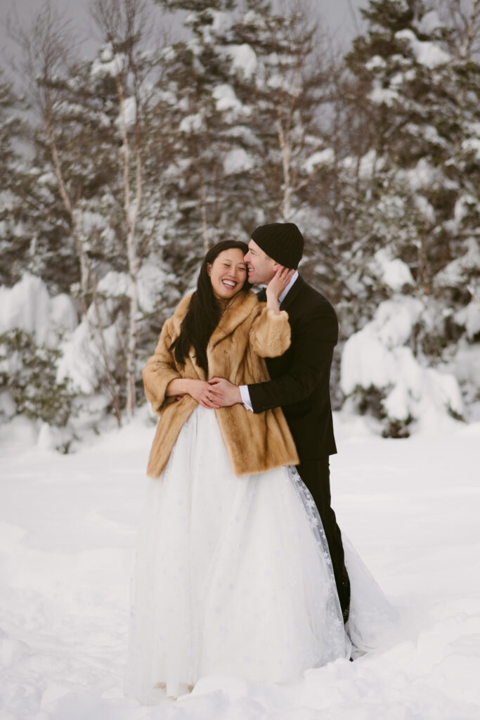 A bride leans against her groom, smiling while touching his cheek during their snowy winter wedding in upstate New York.