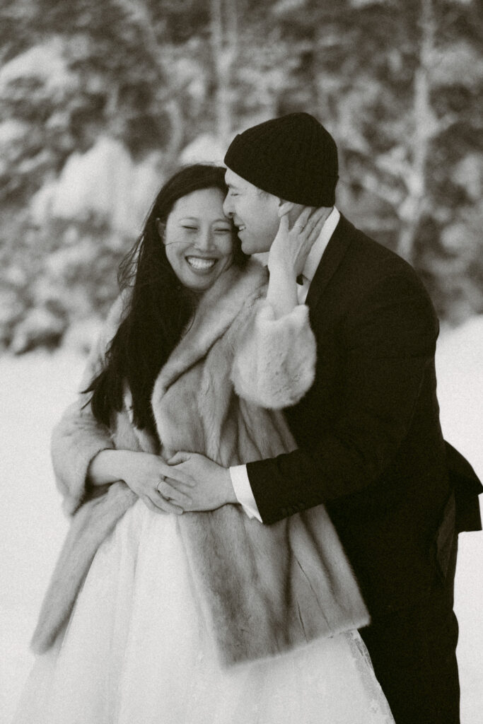 A black and white image of a bride leaning against her groom, smiling while touching his cheek during their snowy winter wedding in upstate New York.