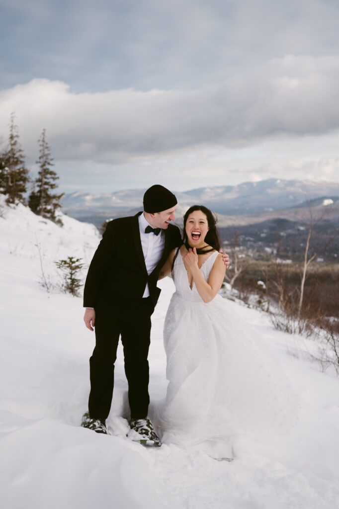 A groom laughs at his bride as she reacts to the cold winter wind during their snowy hiking elopement in Lake Placid, NY.
