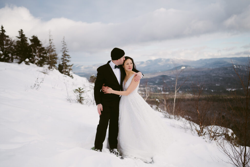 A groom hugs his bride in close and kisses her on the temple while she gazes at Whiteface Mountain during their winter elopement in Lake Placid, NY.