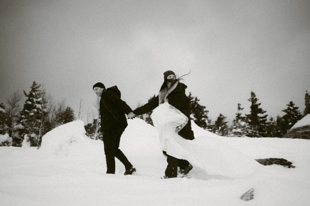 A black and white image of a man leading a woman through deep snow while wearing snowshoes in Lake Placid, NY.