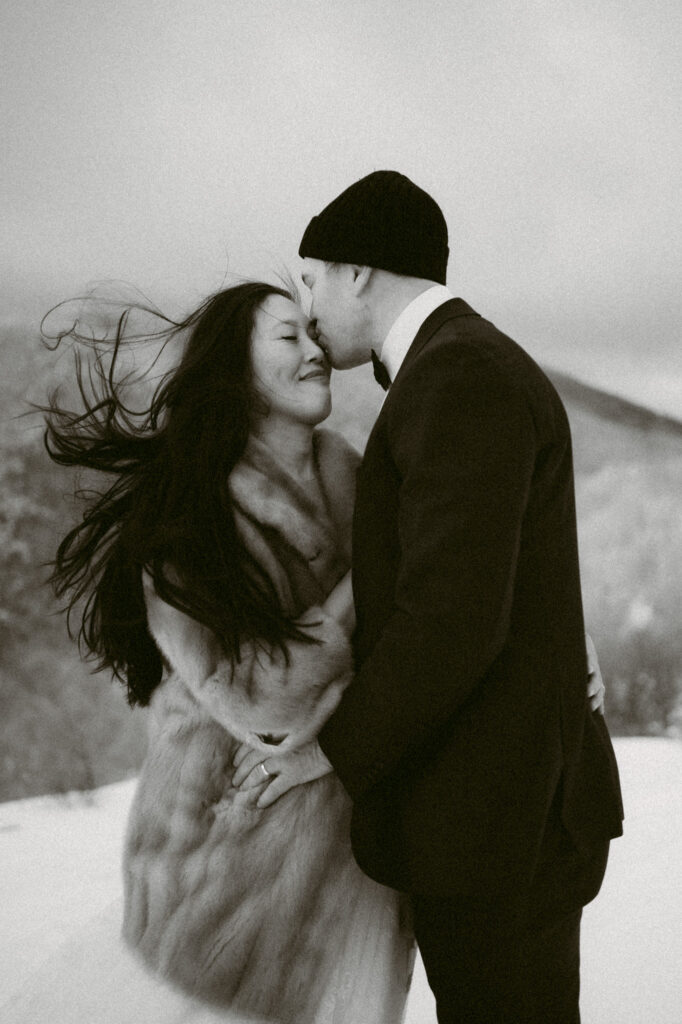A black and white image of a groom kissing his bride on the cheek while her hair blows in the wind during their hiking elopement in upstate New York.