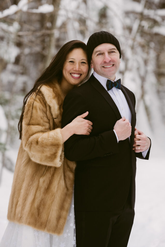 A bride wearing a fur coat hugs her groom while they pose for wedding portraits on a snowy trail.