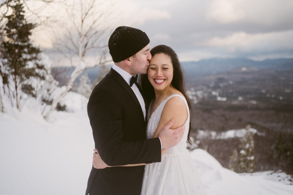 A groom holds his bride while kissing her temple. Lake Placid, NY can be seen off in the distant background.