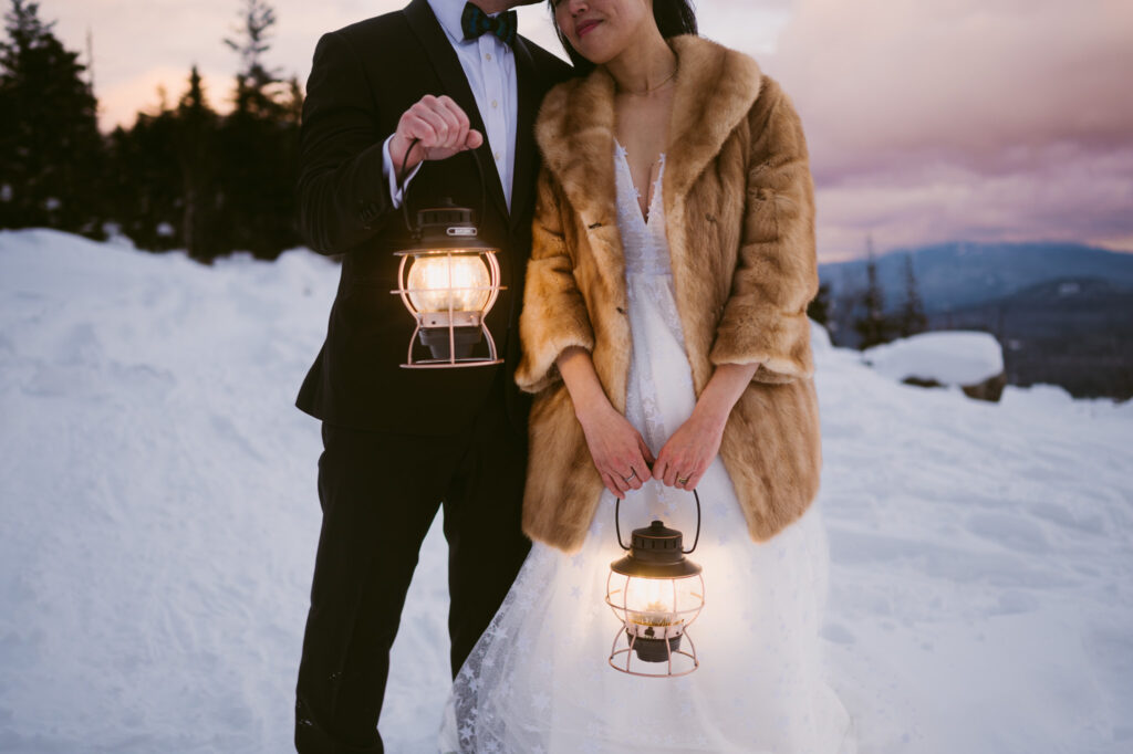 A bride and groom hold lanterns as the sun begins to set during their winter Adirondack elopement.