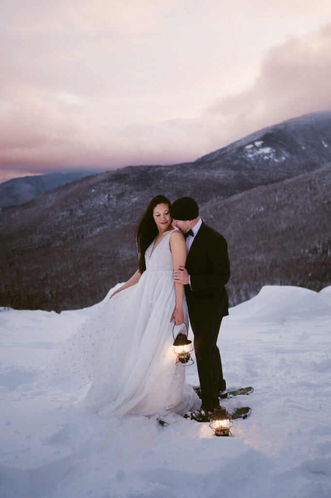 A bride and groom hold lanterns as the sun begins to set during their winter Adirondack elopement. Whiteface Mountain can be seen in the background.