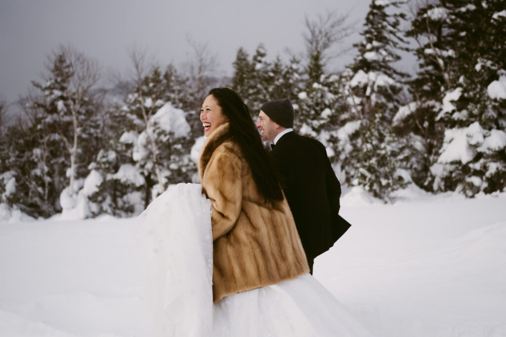 A bride and groom, bundled up and wearing snowshoes, wade through the deep snow during their winter elopement in Lake Placid, New York.