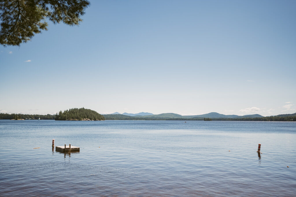 The view of Upper Saranac Lake from the shoreline. An island sits off to the left with the Adirondack Mountains in the background.