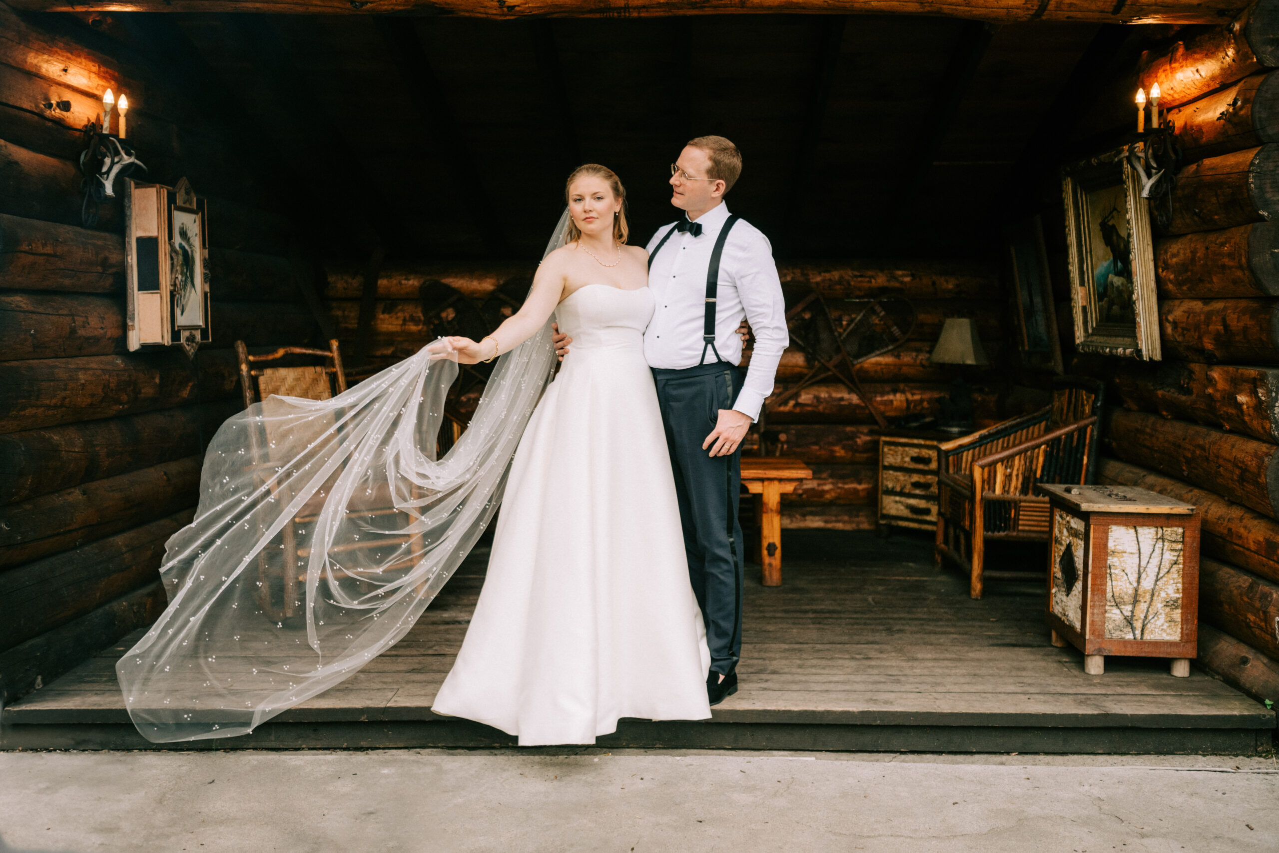 Wedding couple posing on the rustic porch of a log cabin at Whiteface Lodge in the Adirondacks, with the bride showcasing her elegant veil and strapless gown while the groom, dressed in suspenders and a bow tie, looks on lovingly.